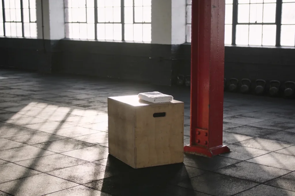 An empty warehouse gym at first light with a timber plyo box topped by a folded white towel, a red steel column and kettlebells along the wall